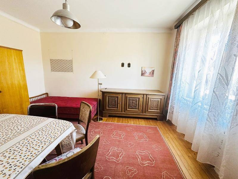 Heller Wohnraum mit Esstisch, Bett und traditionellen Möbeln, gut beleuchtet durch ein großes Fenster.