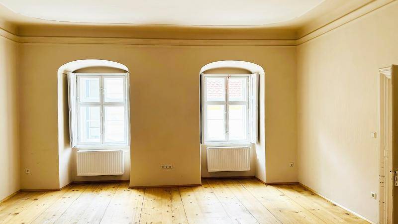 Heller Raum mit Holzdielenboden, hohen Decken und zwei großen Fenstern mit Heizkörpern.
