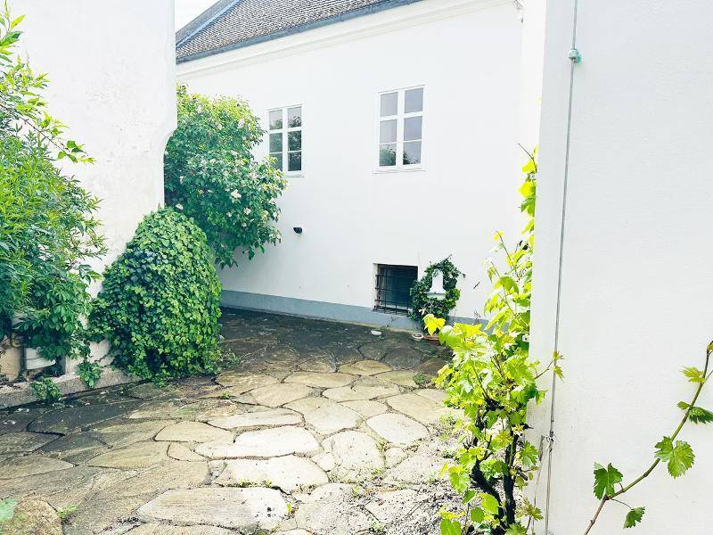 Begrünter Innenhof mit Steinplatten und einer weißen Gebäudefassade unter hellem Himmel.