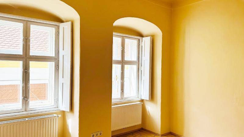 Heller Raum mit gelben Wänden, Holzdielenboden und zwei großen Fenstern mit Fensterläden.