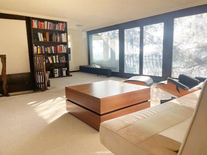 Geräumiges Wohnzimmer mit Teppichboden, Bücherregal und großen Fenstern mit Blick ins Grüne.