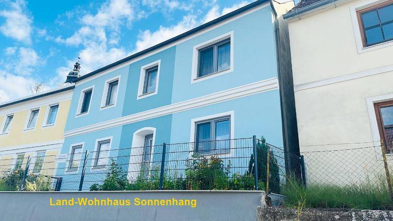 Seitenansicht des Land-Wohnhauses mit blauer Fassade und angrenzendem Gebäude unter klarem Himmel.
