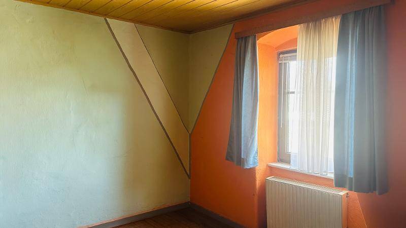Helles Zimmer mit Holzbalkendecke, farbigen Wänden und einem Fenster mit Heizkörper darunter.