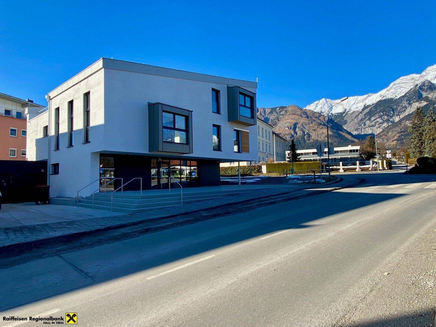 Modernes Gebäude mit weißer Fassade und großen Fenstern, umgeben von einer Berglandschaft unter blauem Himmel.