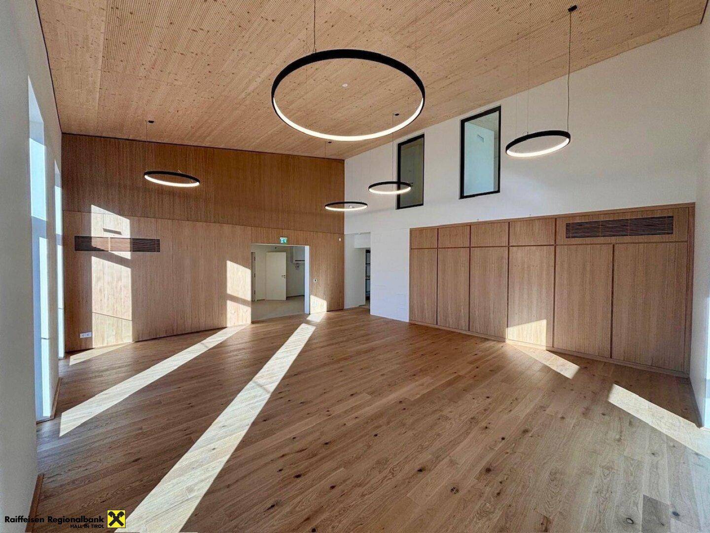 Großer, heller Raum mit Holzboden und Holzverkleidung an Wänden und Decke, ausgestattet mit modernen Ringlampen.