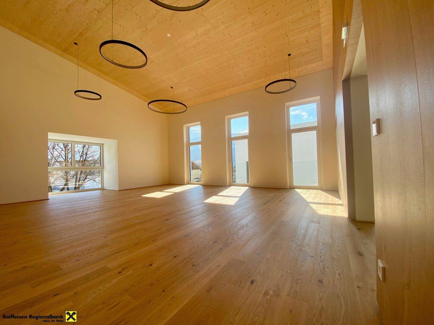 Großer, heller Mehrzweckraum mit Holzboden und hohen Decken, durchflutet von natürlichem Licht.