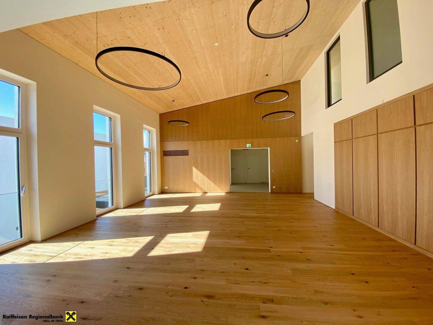Heller Mehrzweckraum mit schrägen Holzdecken, großen Fenstern und integrierten Holzschränken.