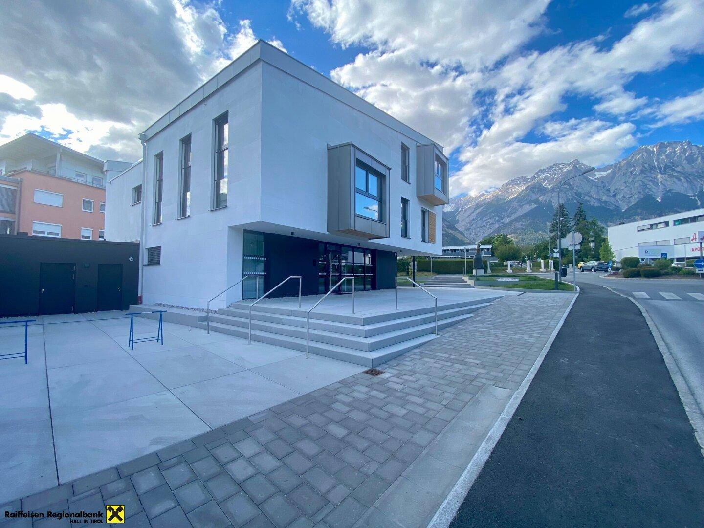 Modernes Geschäftsgebäude mit breiter Eingangstreppe und beeindruckendem Bergblick im Hintergrund unter bewölktem Himmel.