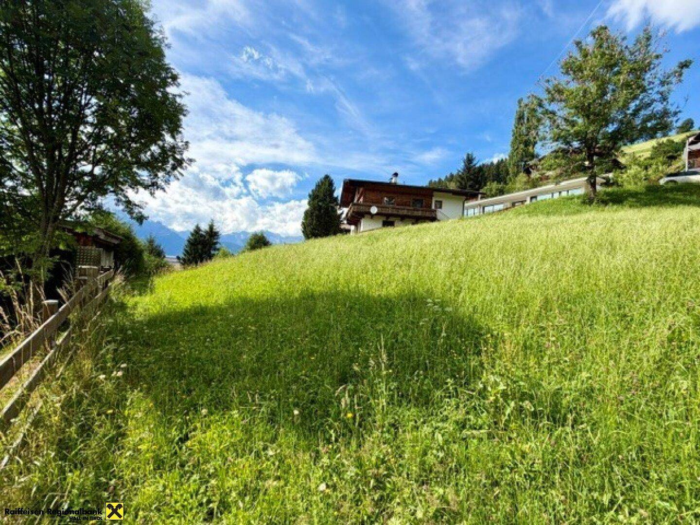 Weitläufige grüne Wiese mit einem Haus im Hintergrund und Blick auf die Alpen.