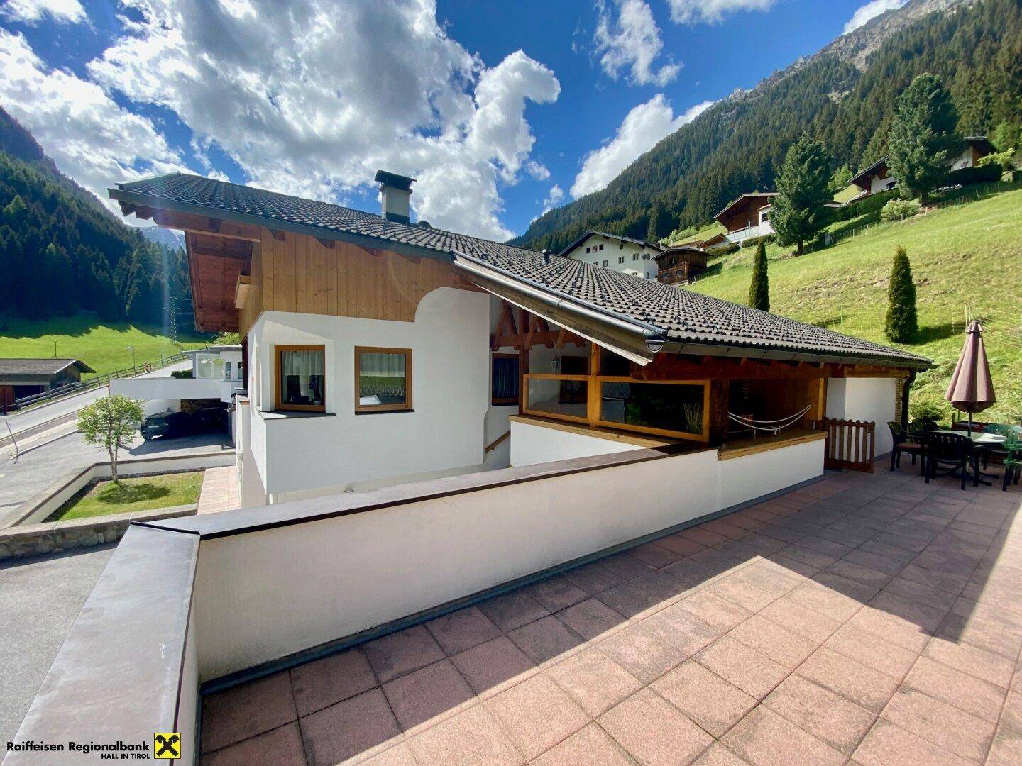 Hausfassade mit großer gepflasterter Terrasse und Bergblick in alpiner Umgebung.