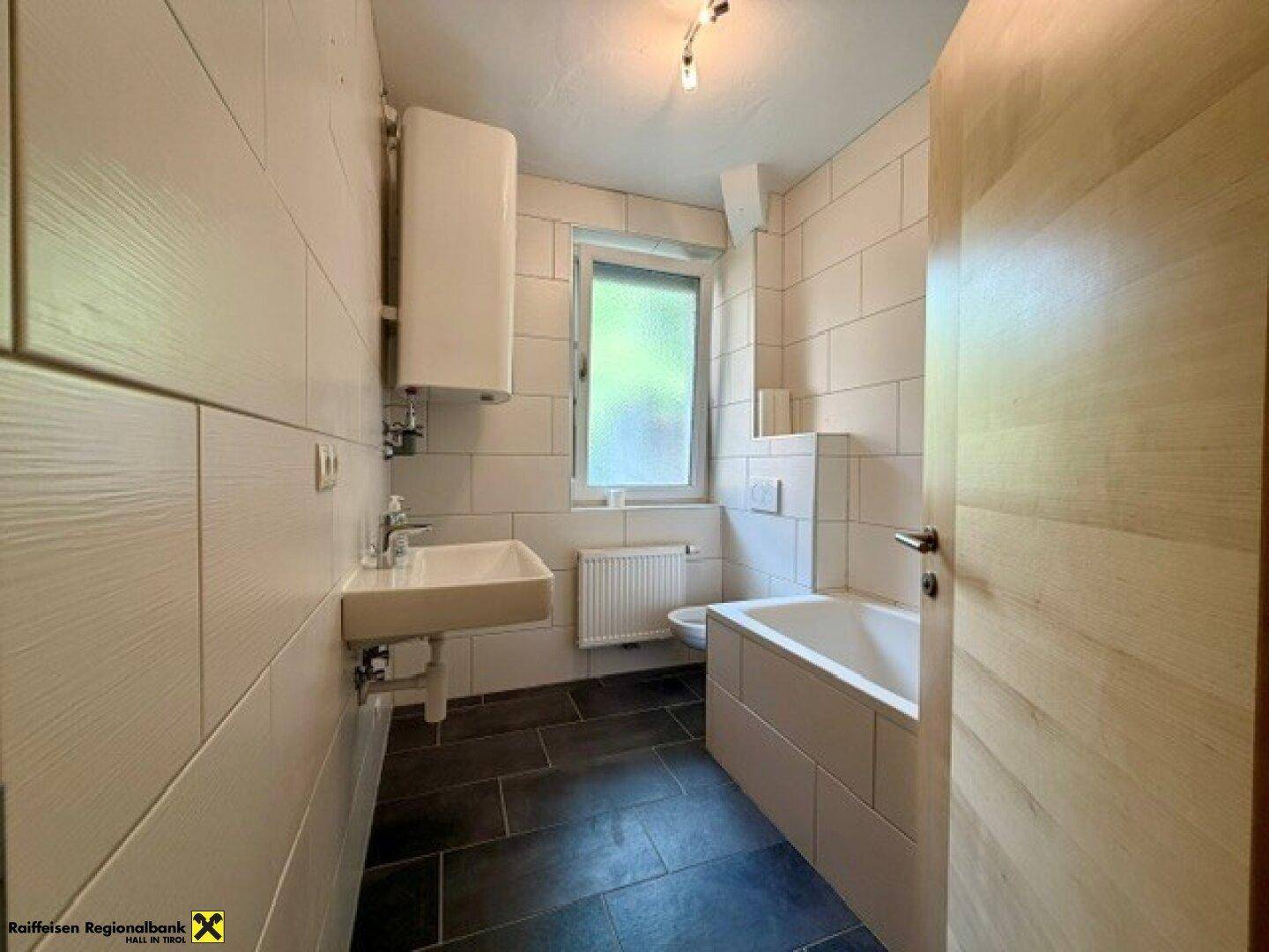 Modern gefliestes Badezimmer mit Badewanne, Waschbecken und Fenster für Tageslicht.
