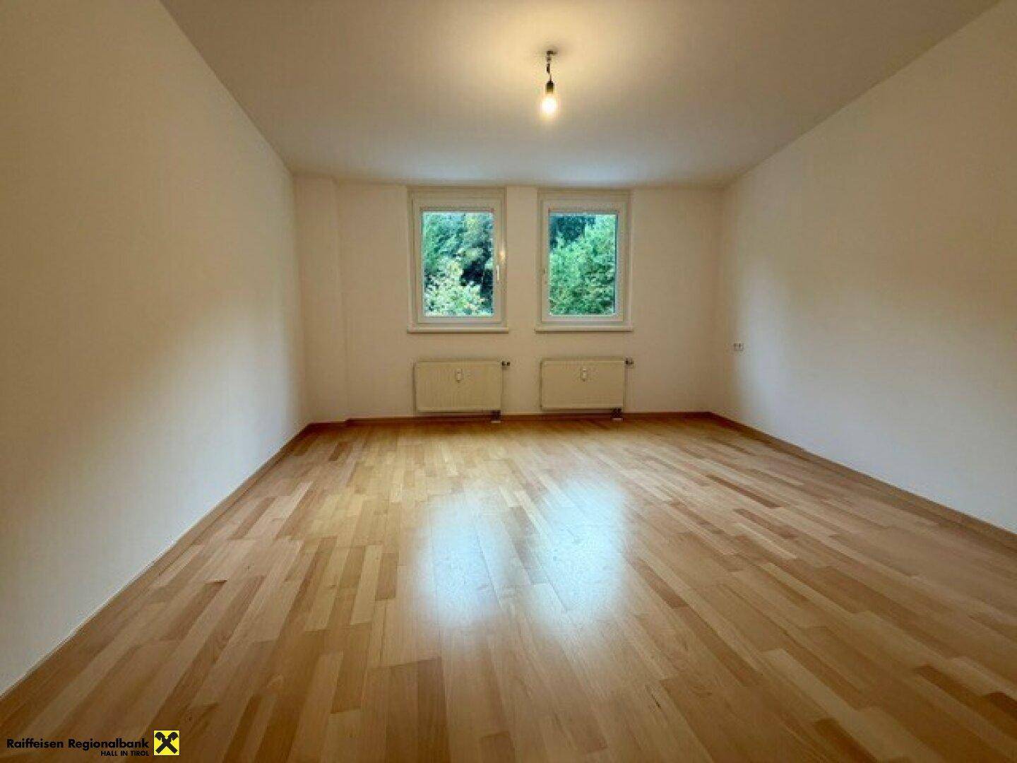 Helles Schlafzimmer mit Holzboden und zwei Fenstern, die einen Blick ins Grüne ermöglichen.