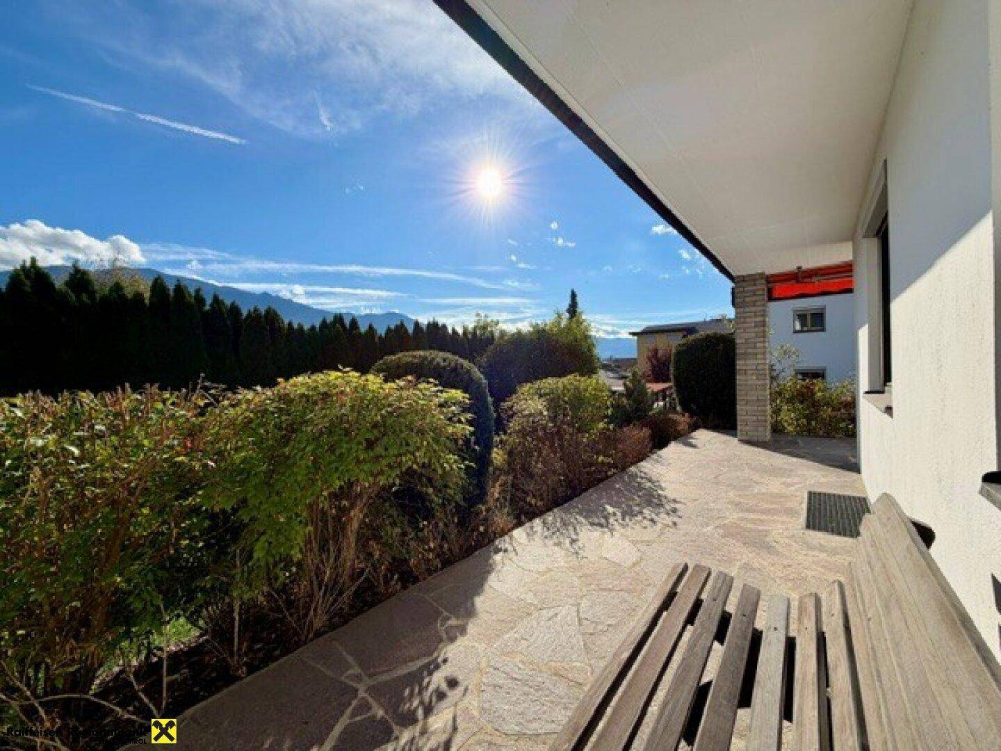 Sonnige Terrasse mit Holzbank, Steinboden und weitem Blick auf die umliegende Berglandschaft und Natur.