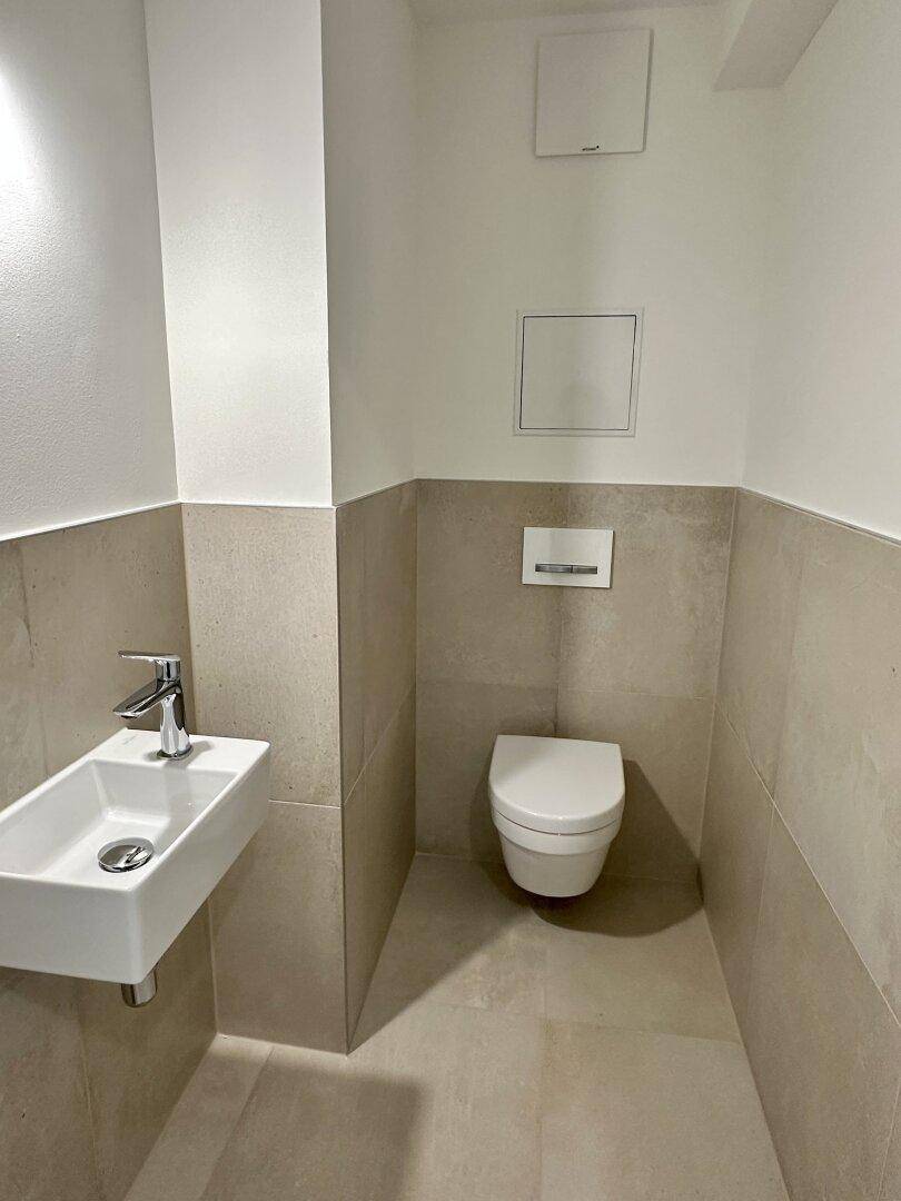 Modernes Gäste-WC mit wandhängender Toilette und kleinem Waschbecken, hell gefliest.