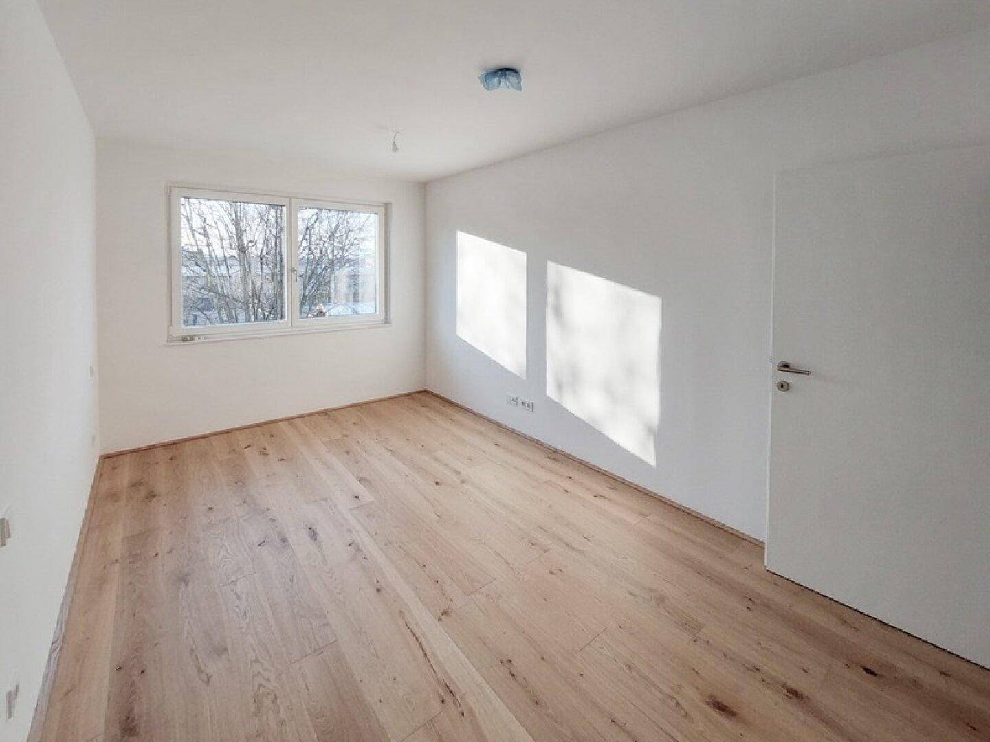 Helles Schlafzimmer mit neuem Holzparkettboden und großem Fenster, das viel Tageslicht hereinlässt.