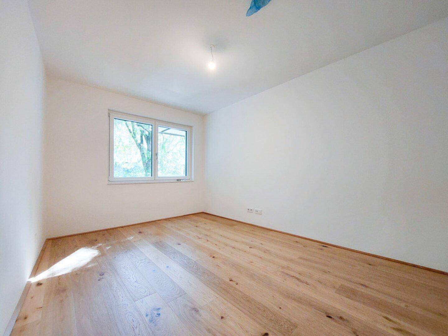 Helles Schlafzimmer mit Holzplankenboden und Fenster mit Blick ins Grüne.