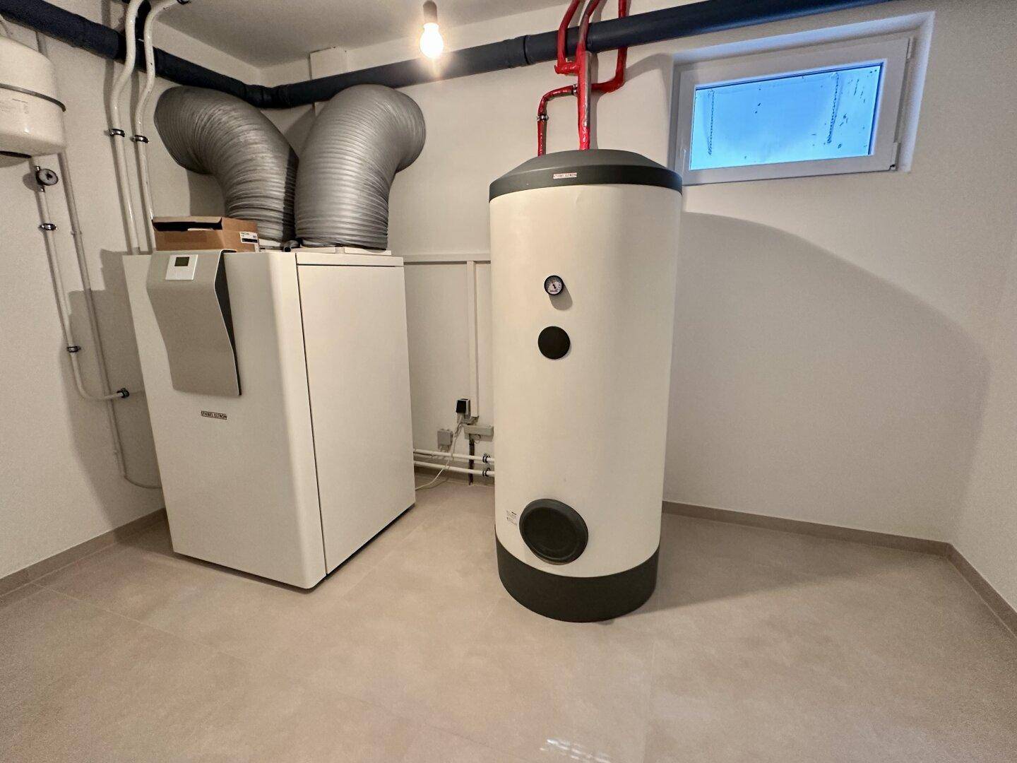 Technikraum im Keller mit moderner Heizungsanlage, Warmwasserspeicher und kleinem Fenster.