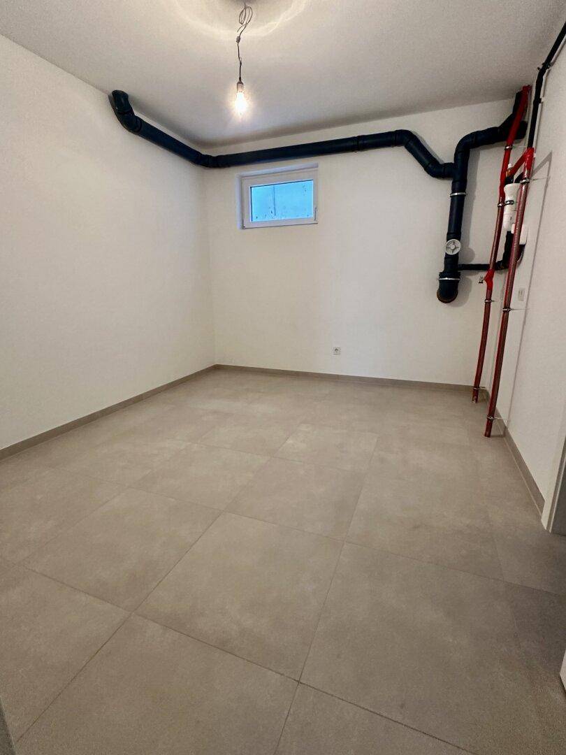 Geräumiger Kellerraum mit Fliesenboden, sichtbaren Rohren und einem kleinen Fenster für Belüftung.