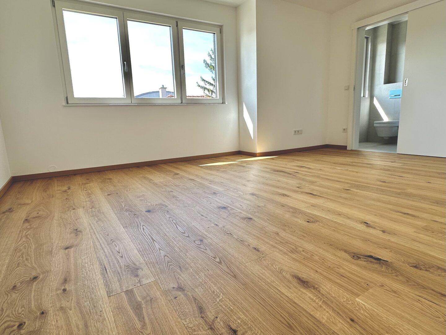 Helles Zimmer mit Holzfußboden und großen Fenstern, mit direktem Zugang zu einem modernen Badezimmer.