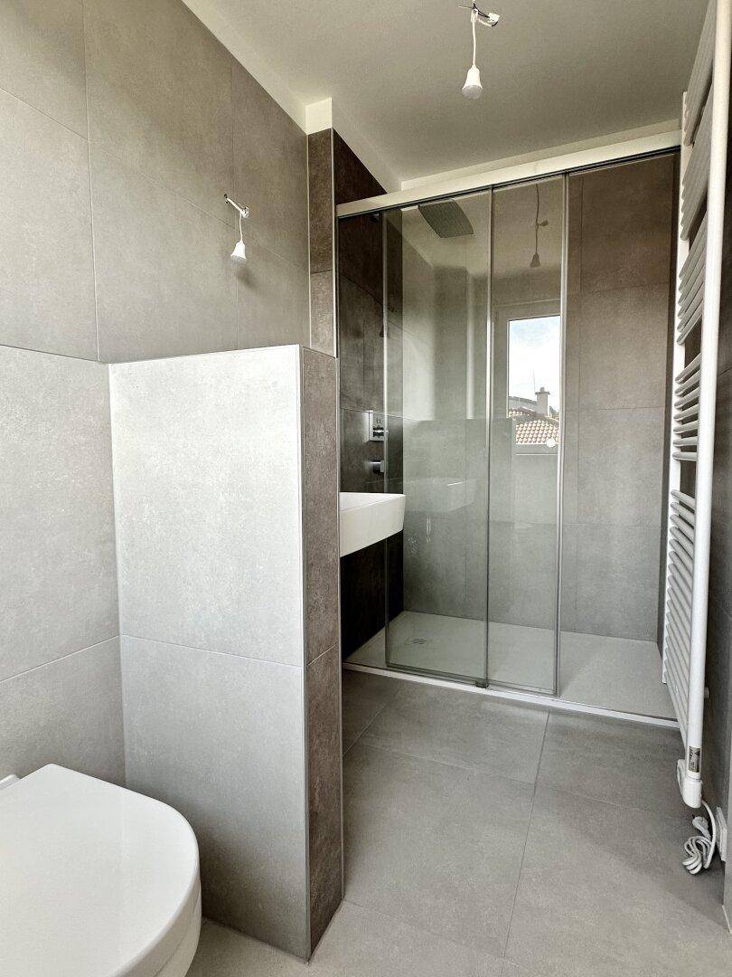 Modernes Badezimmer mit grauen Fliesen, Duschkabine, Wand-WC und beheiztem Handtuchhalter.