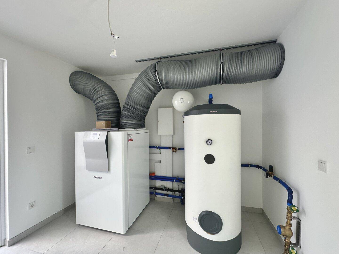 Technikraum mit moderner Heizungsanlage, Warmwasserspeicher und Lüftungssystem auf Fliesenboden.