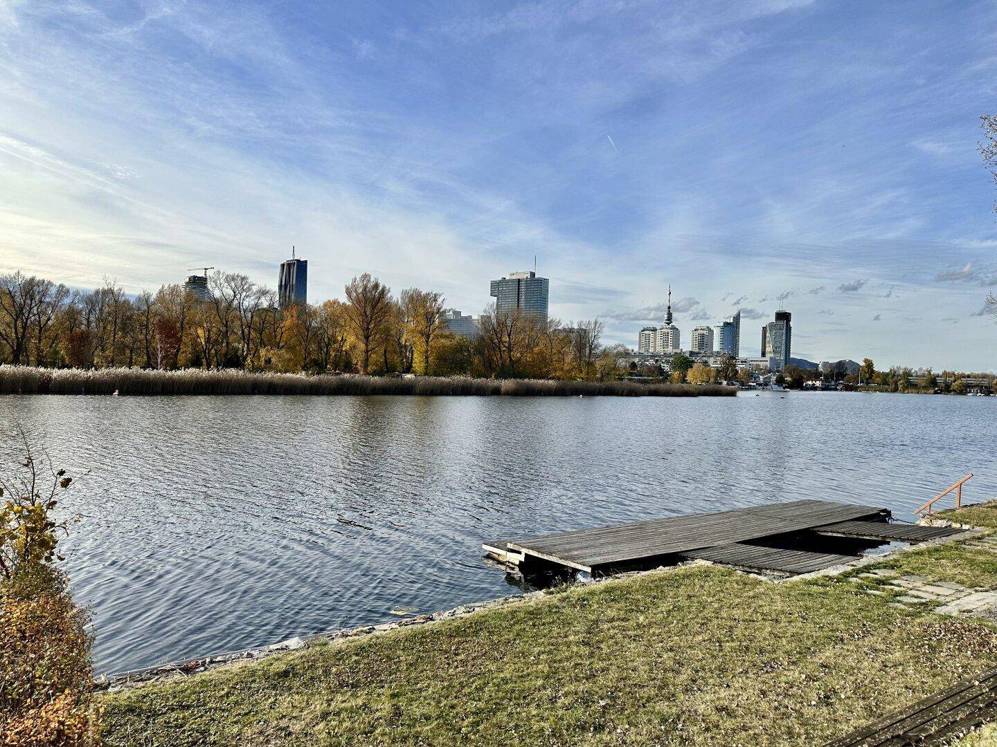 Weitläufiger Blick über einen See mit einem Steg und der Skyline einer Stadt im Hintergrund.