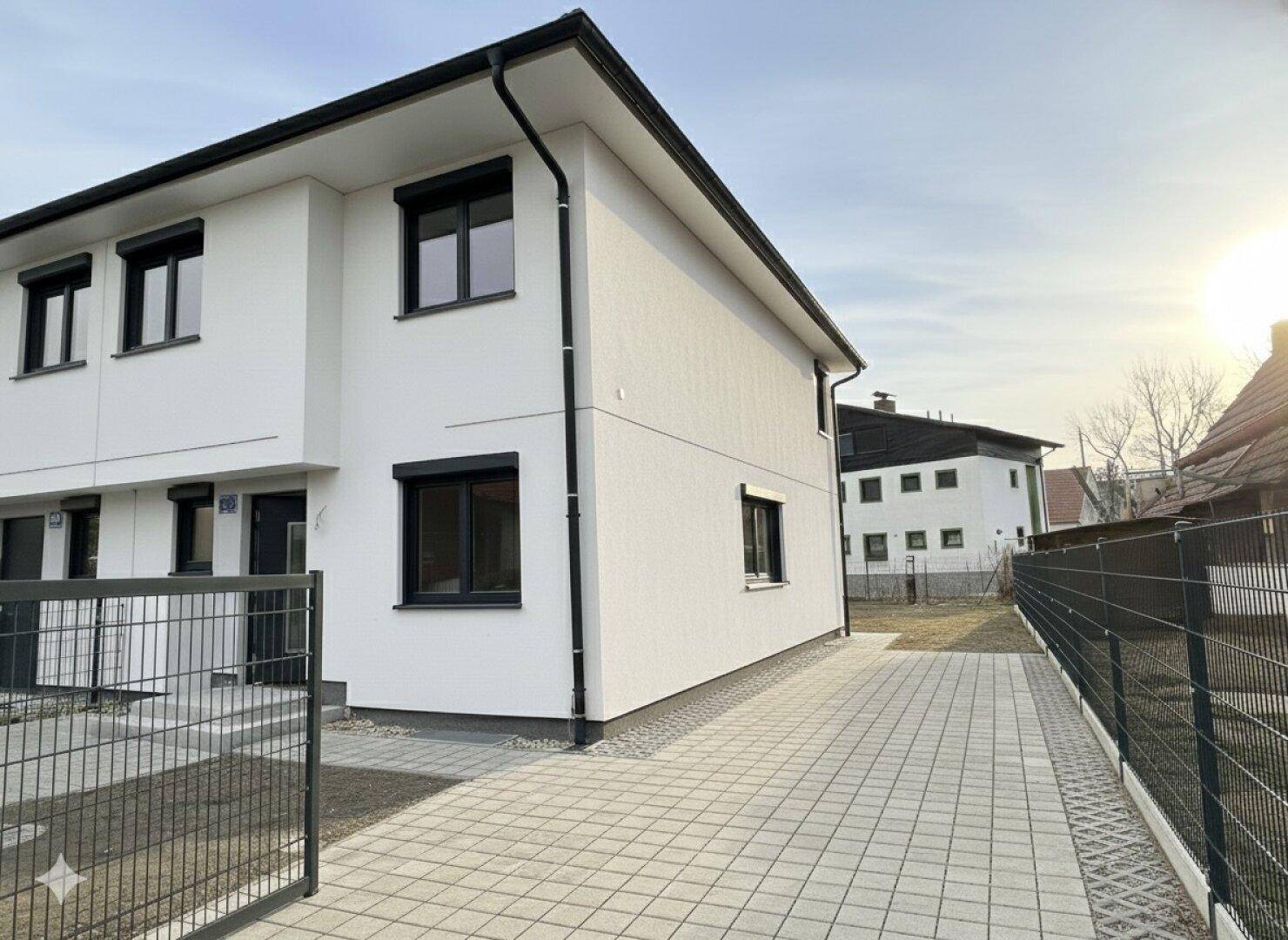 Moderne weiße Hausfassade mit gepflasterter Einfahrt und schwarzem Zaun unter blauem Himmel.