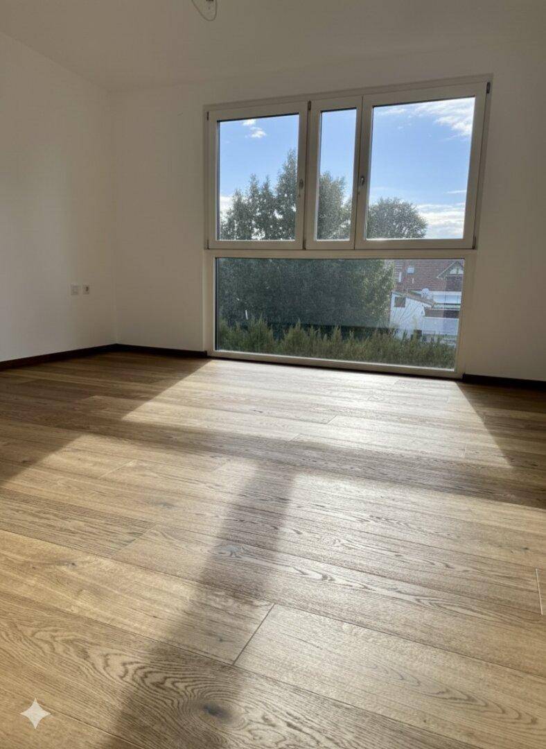 Lichtdurchflutetes Zimmer mit großem Fenster und Blick ins Grüne, ausgestattet mit Holzfußboden.