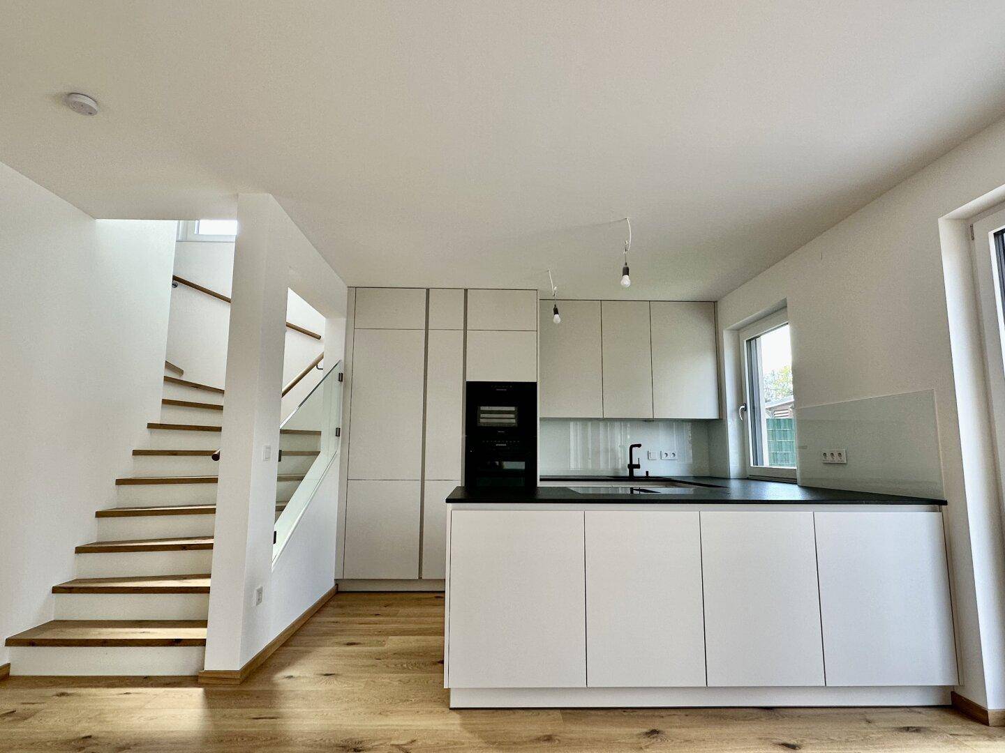 Moderne Einbauküche mit weißen Schränken, schwarzer Arbeitsplatte und integriertem Backofen neben der Holztreppe.