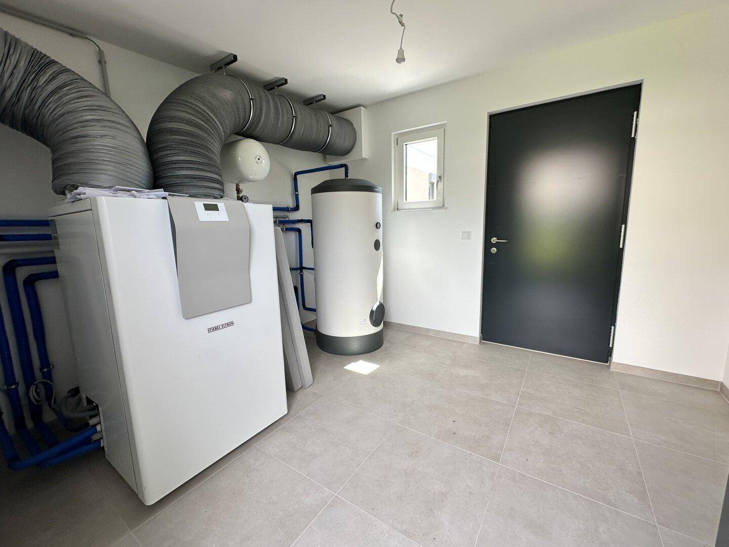 Technikraum mit Heizungsanlage, Warmwasserspeicher und Lüftungsrohren, gefliestem Boden und Fenster.