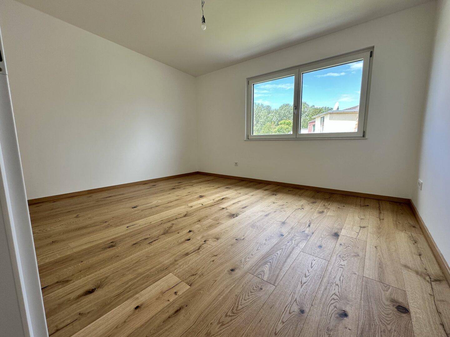 Helles Schlafzimmer mit Holzboden und großem Fenster, das viel Tageslicht hereinlässt.