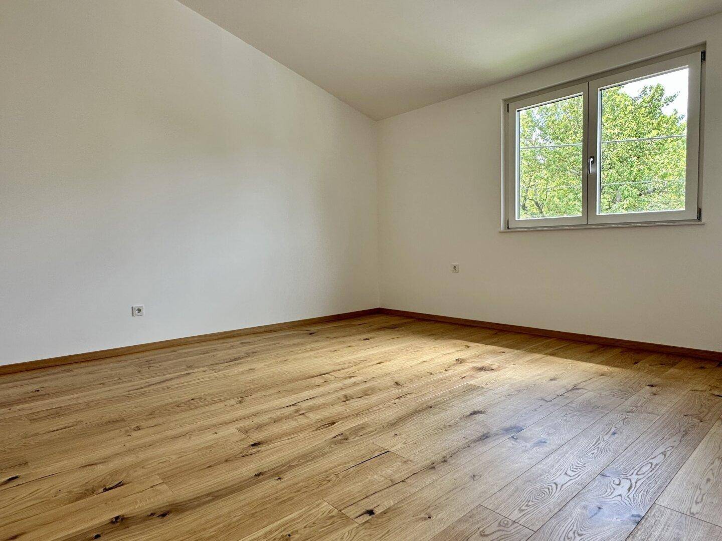 Leeres Schlafzimmer mit Holzboden und Fenster mit Blick ins Grüne, bereit zur individuellen Gestaltung.