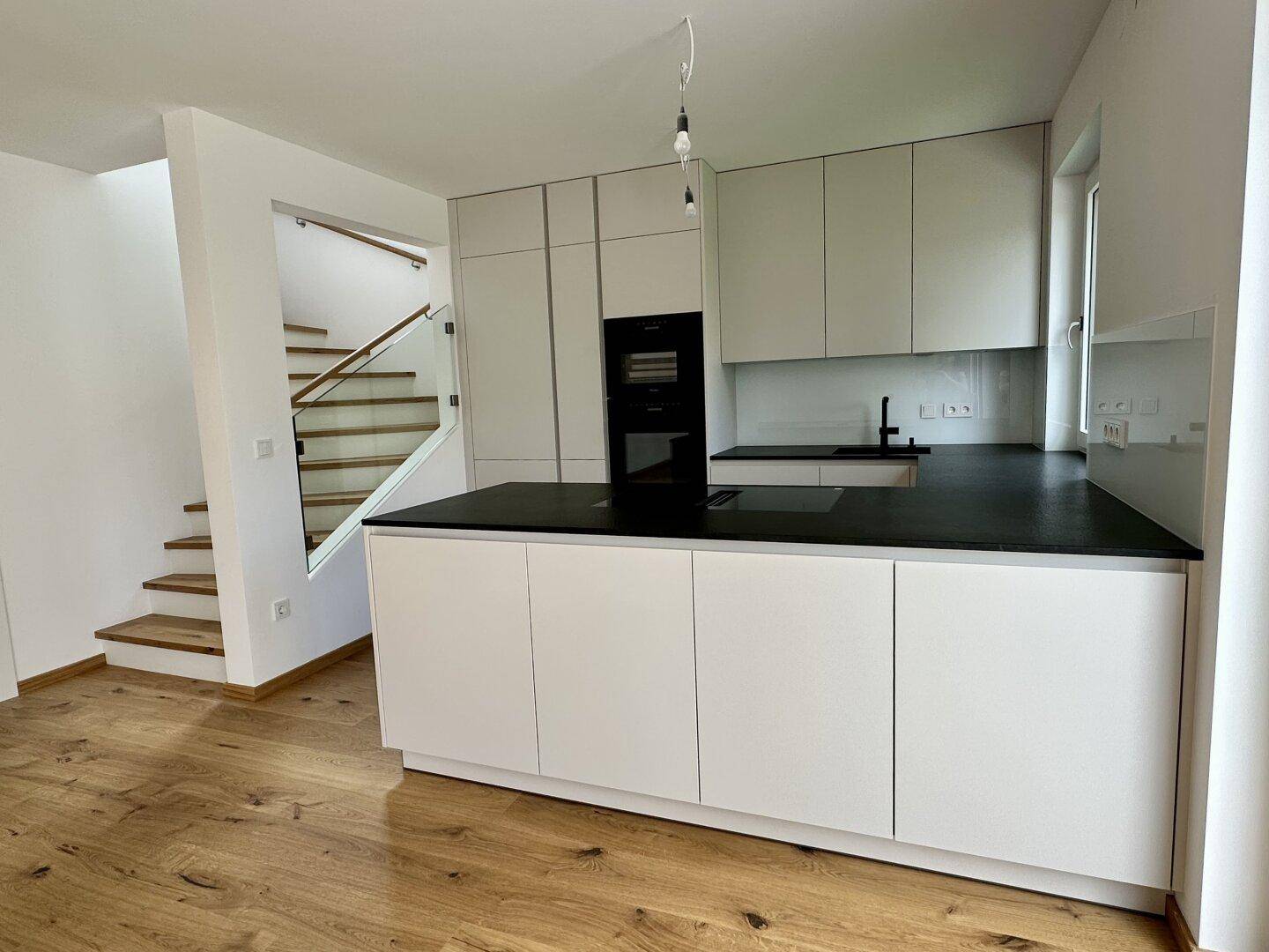 Moderne Einbauküche mit weißen Schränken, schwarzer Arbeitsplatte und integriertem Backofen neben der Holztreppe.