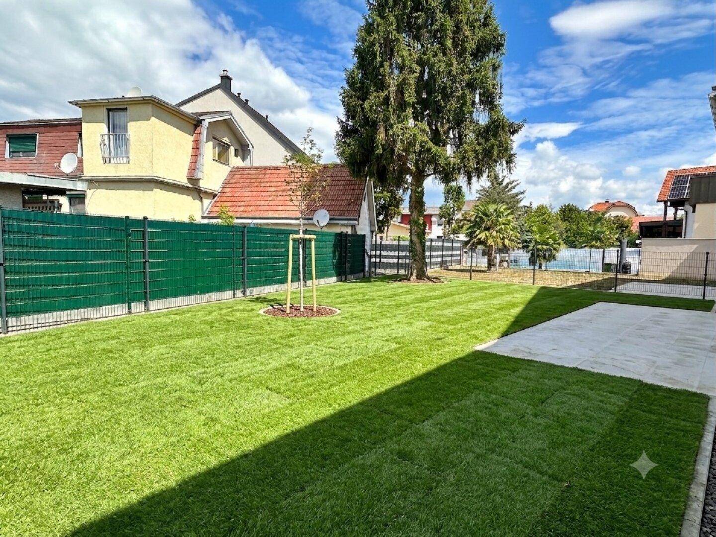 Gepflegter Garten mit frischem Rasen, jungem Baum und Blick auf die umliegenden Häuser.