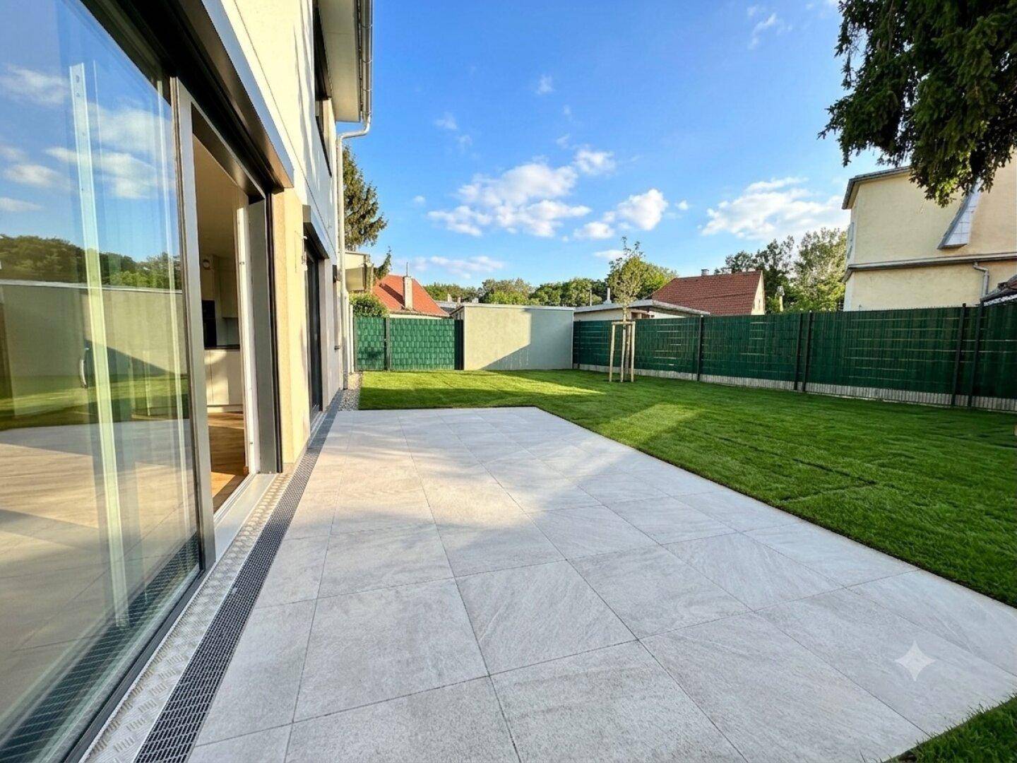 Gepflasterte Terrasse mit angrenzendem Rasen und großen Glasschiebetüren zum Wohnbereich.