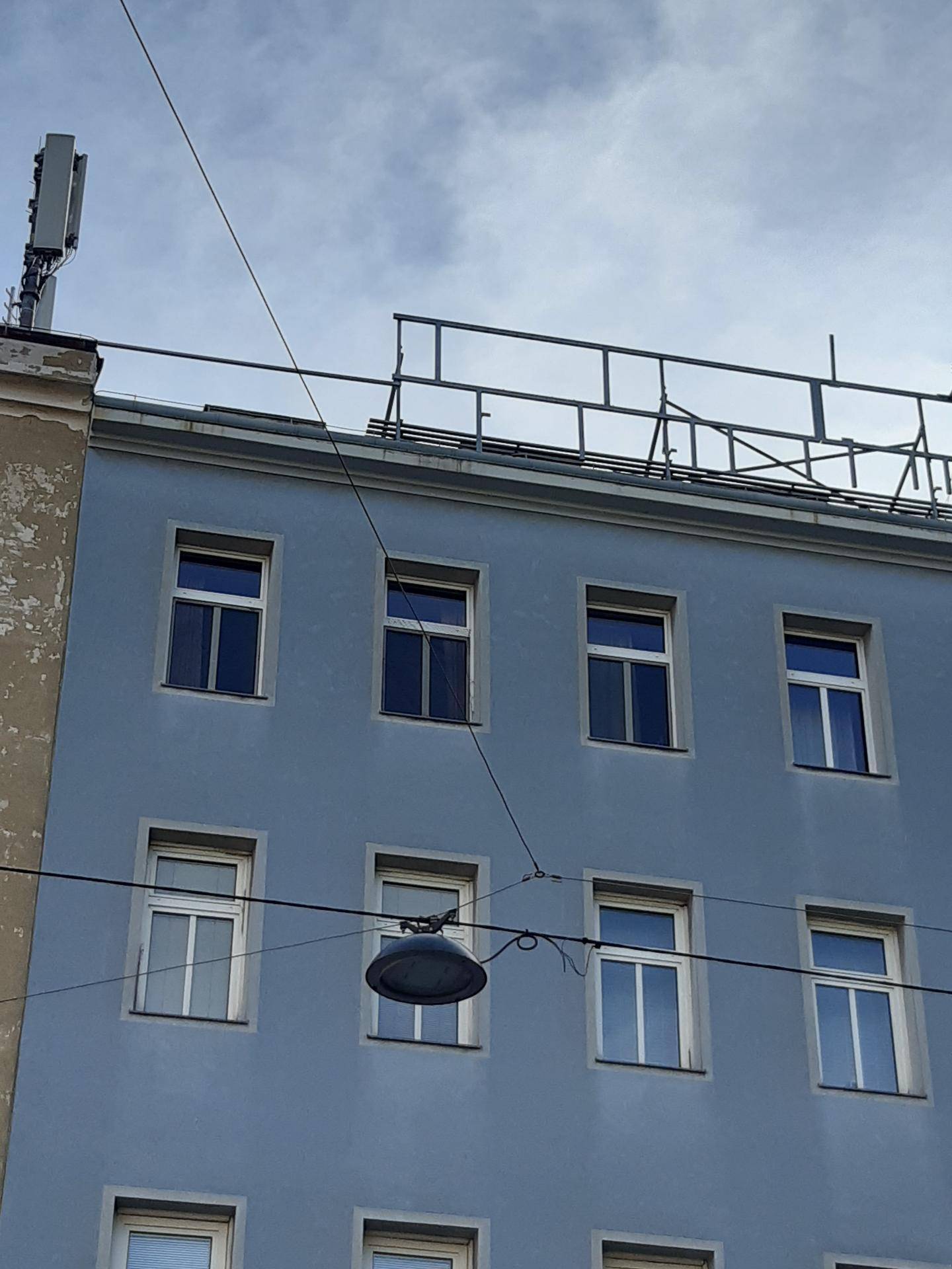 Oberer Teil der blauen Gebäudefassade mit Fenstern und Dachkonstruktion unter bewölktem Himmel.