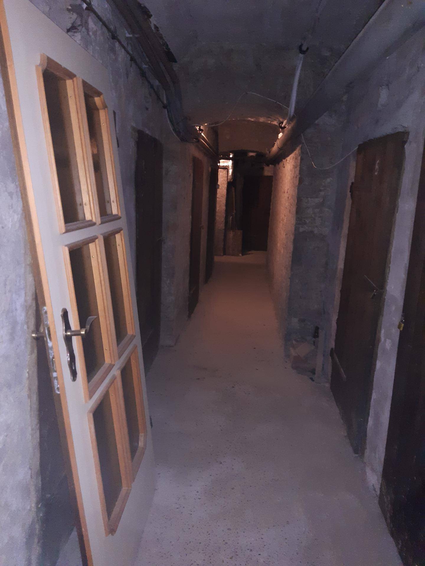 Langer, schmaler Kellergang mit mehreren Lagerräumen und einfacher Beleuchtung.
