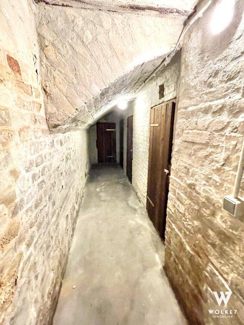 Langer, schmaler Kellergang mit unverputzten Ziegelwänden und Holztüren zu Abstellräumen.