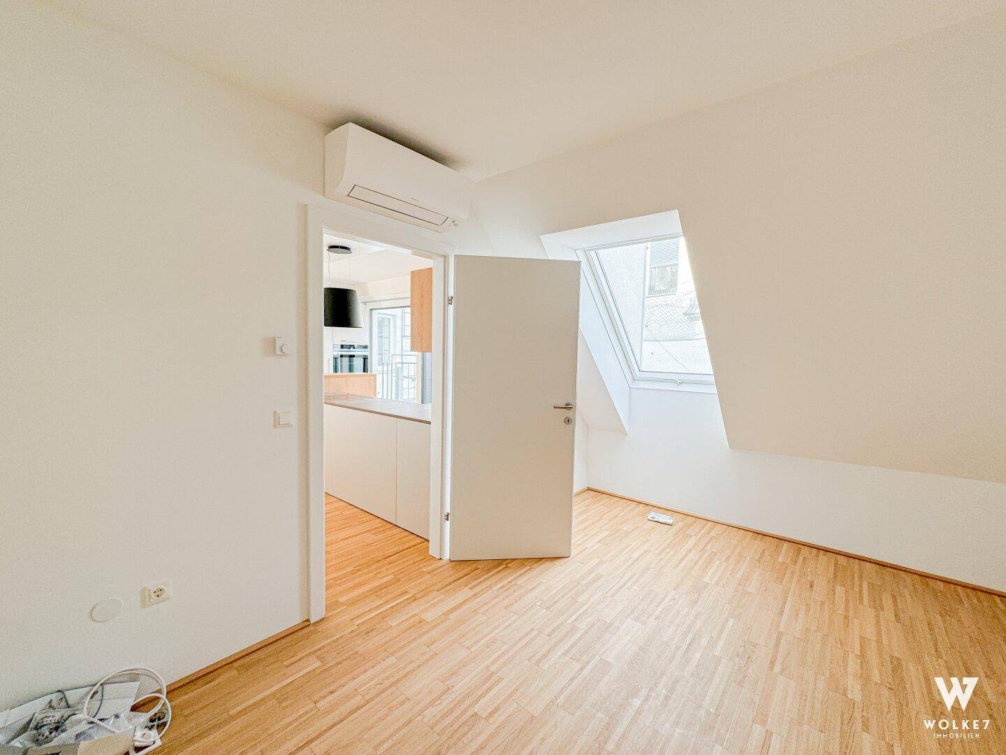 Helles Zimmer mit Holzboden, Dachfenster und Klimaanlage, mit Blick zur Küche.