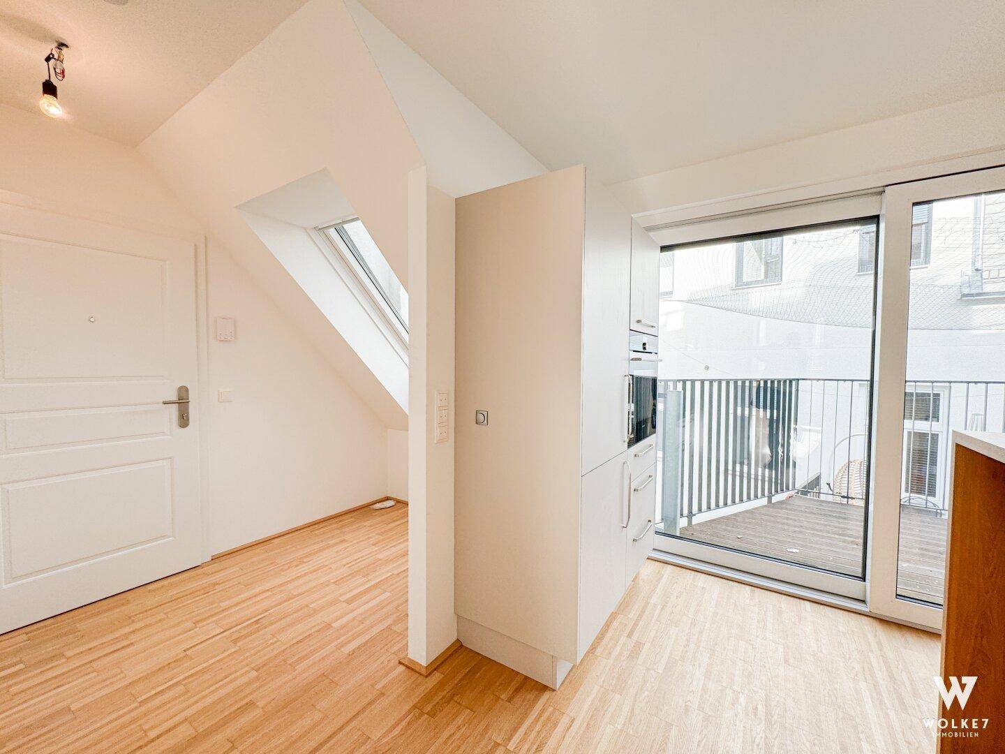 Offener Bereich mit integrierter Küchenzeile, Holzboden und Zugang zu einem Balkon.