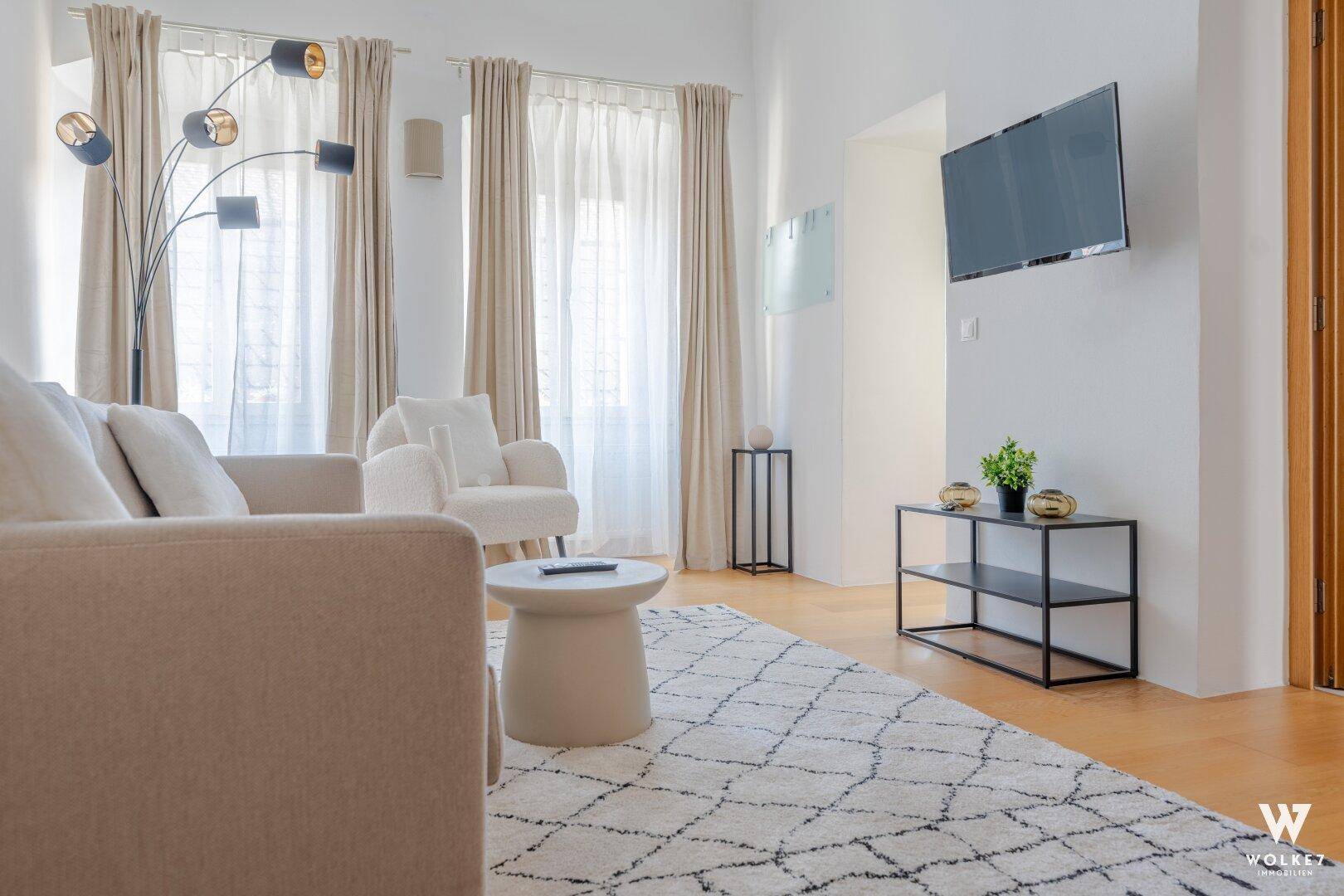 Modern eingerichtetes Wohnzimmer mit Wandfernseher, beigem Sofa und stilvollem Teppich.