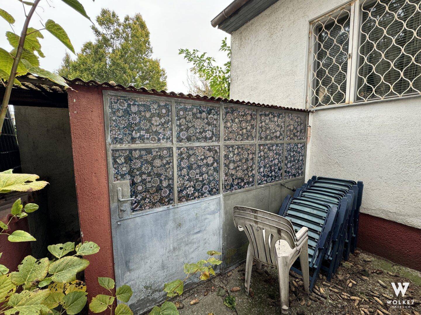 Seitenansicht eines Schuppens mit gemusterten Fenstern und gestapelten Gartenstühlen.