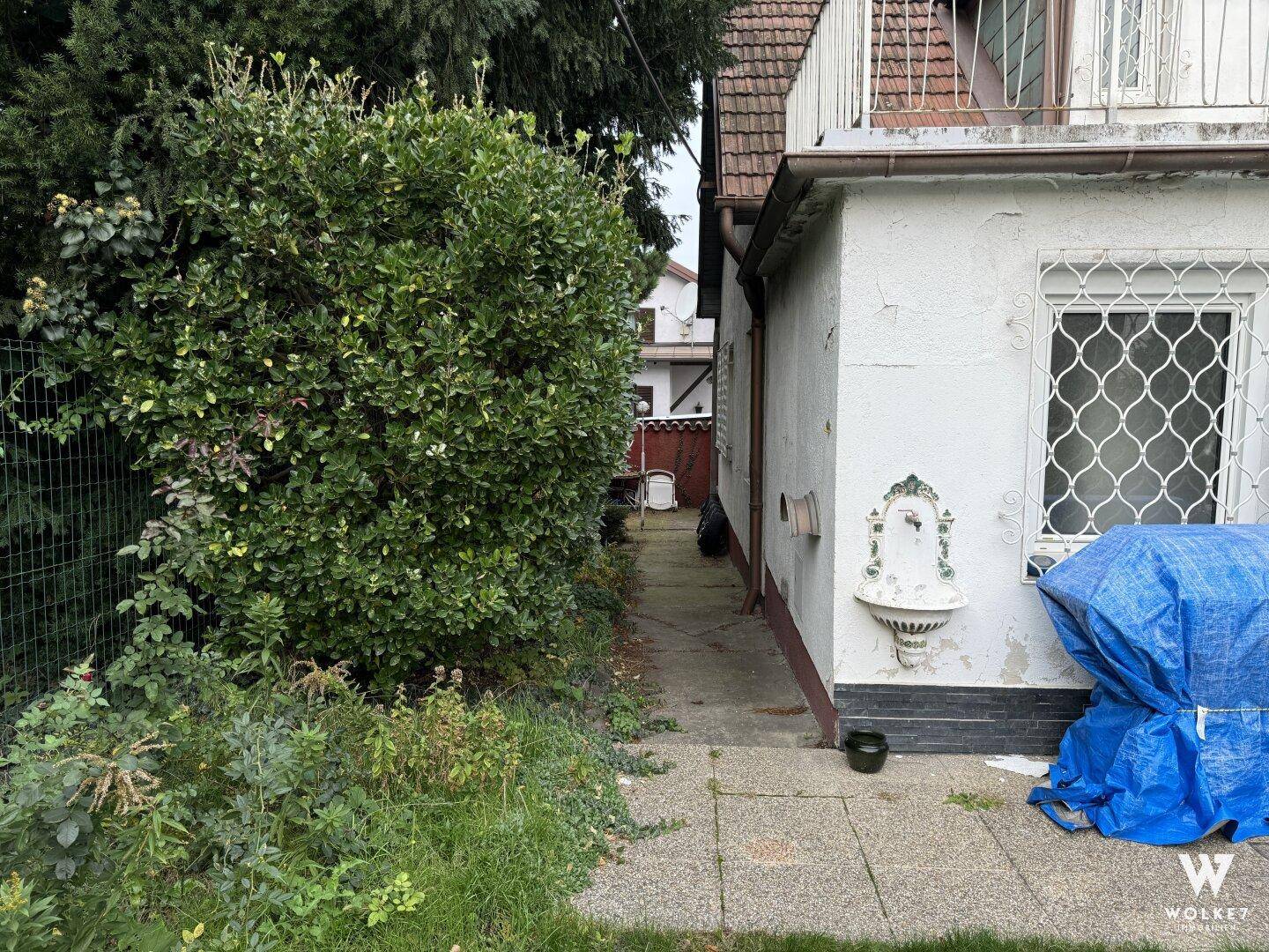 Gartenweg neben dem Haus mit einer dekorativen Wandfontäne und Hecken.