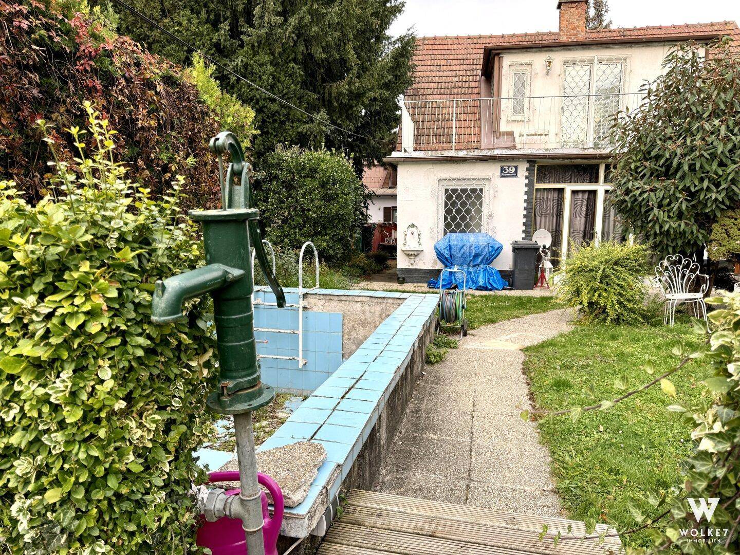 Gartenbereich mit einer alten Wasserpumpe, einem kleinen Becken und einem Weg zum Haus.