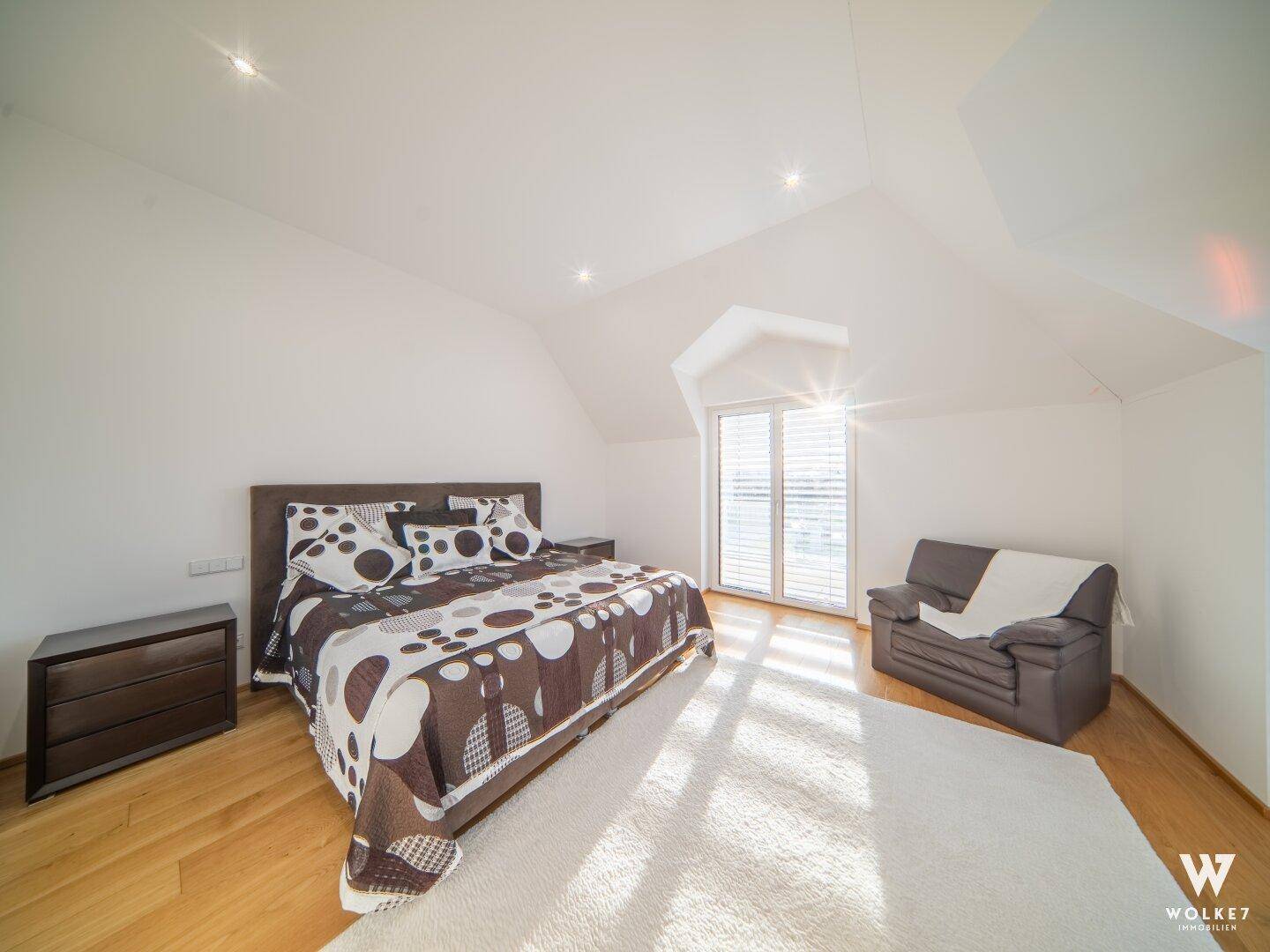 Helles Schlafzimmer mit Holzboden, Dachschrägen und viel Sonnenlicht durch die Fenster.