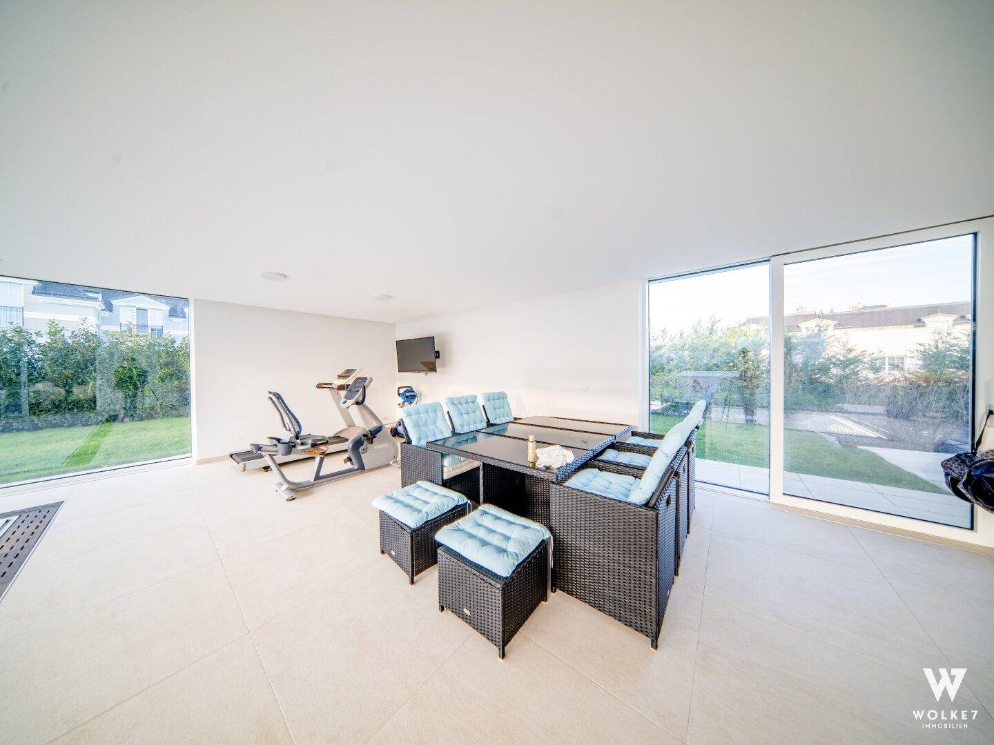Geräumiger Fitnessbereich mit Trainingsgeräten, Lounge-Möbeln und großen Fenstern mit Gartenblick.