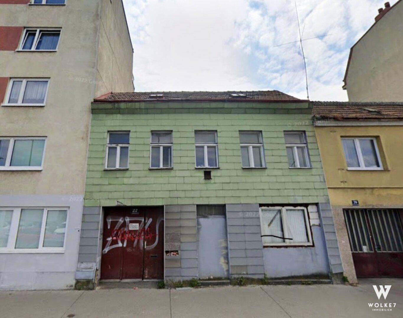 Altes Gebäude mit grüner Fassade und Graffiti an der Straßenseite, umgeben von anderen Häusern.