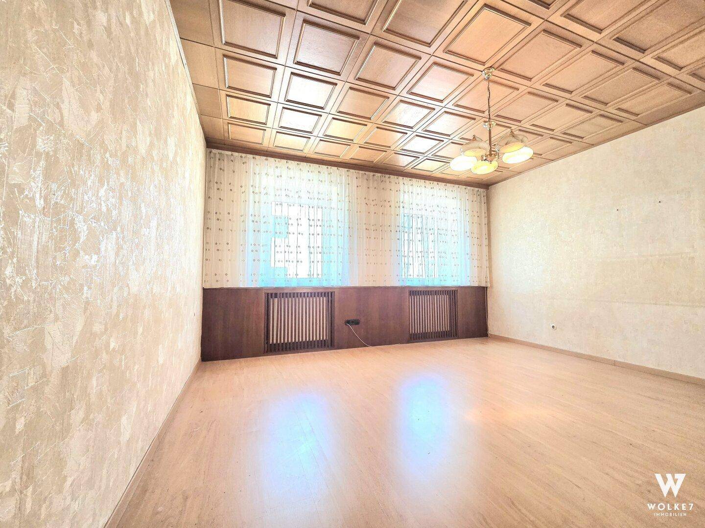 Geräumiger Wohnraum mit Holzpaneeldecke, Parkettboden und zwei Fenstern.