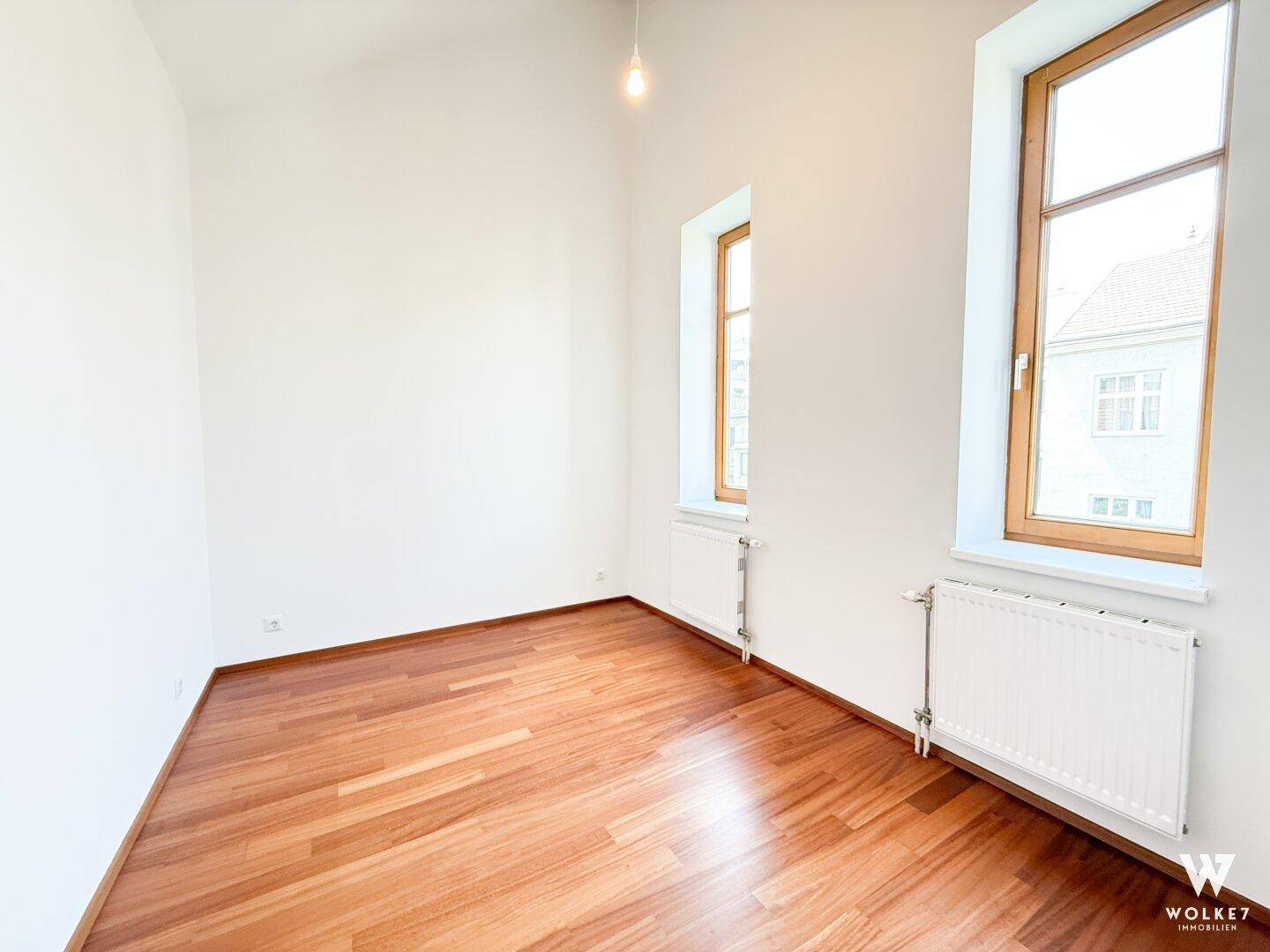 Helles, leeres Zimmer mit Holzboden und großen Fenstern, bereit zur individuellen Gestaltung.