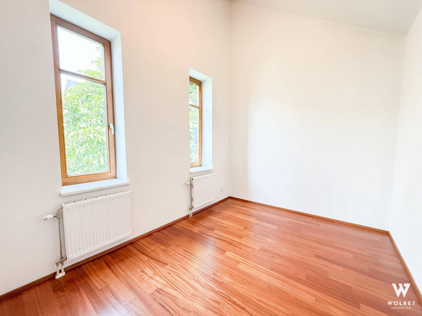 Lichtdurchflutetes Zimmer mit Holzboden und Blick ins Grüne durch die Fenster.